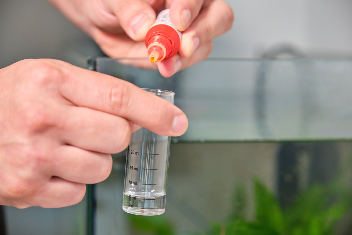 Hands adding test reagent to a water sample beside a freshwater aquarium.