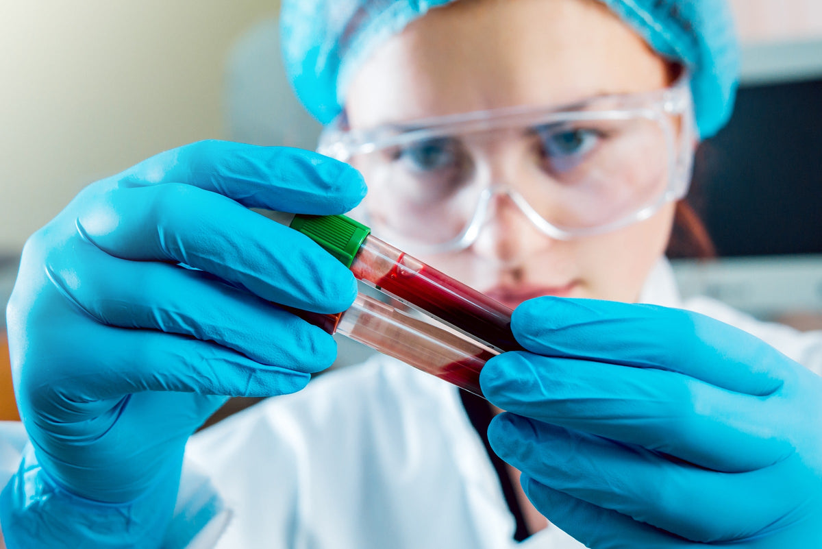 Laboratory worker wearing blue gloves and safety goggles holds two blood sample tubes.