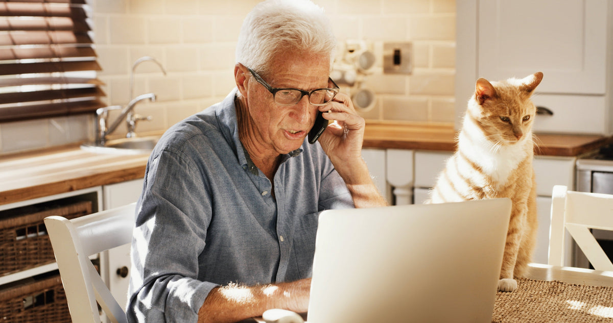 An older person uses a phone and laptop at a kitchen table beside an orange-and-white cat.
