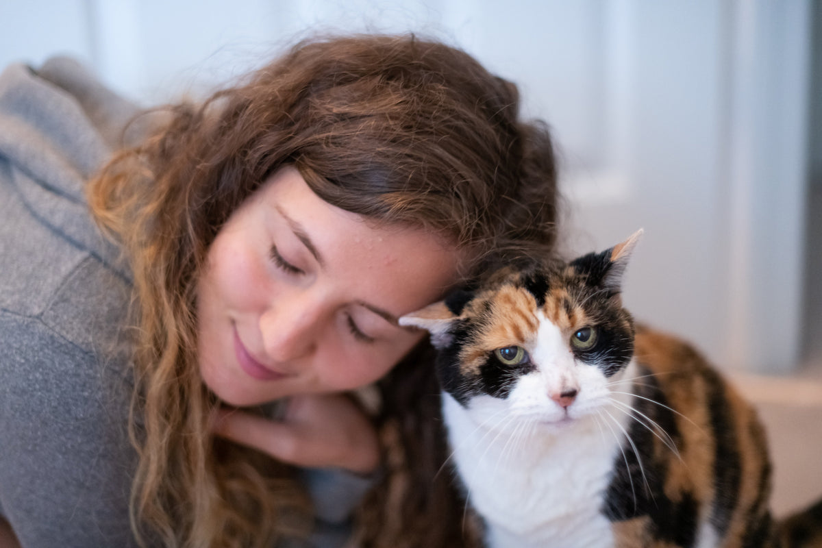 Woman resting her head beside a calico cat on a sofa.