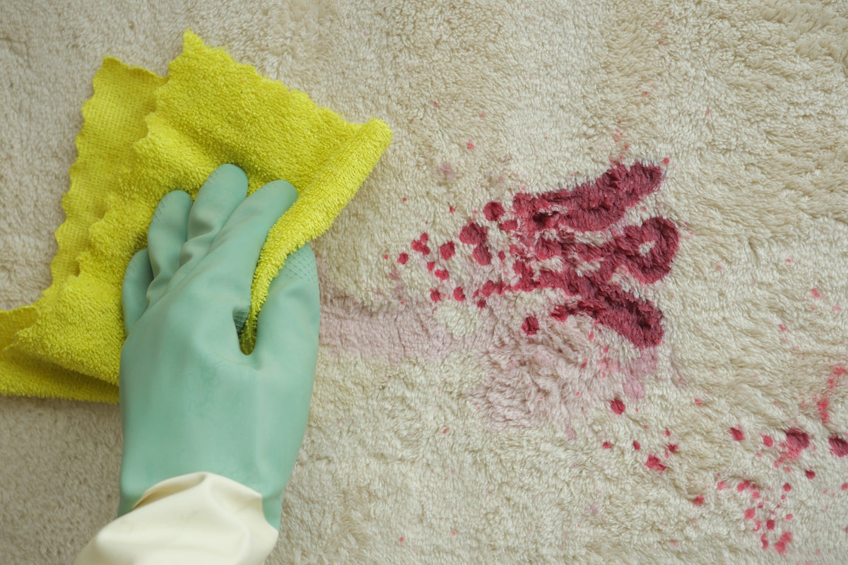 Gloved hand using a yellow cloth to clean a red stain from a pale carpet.