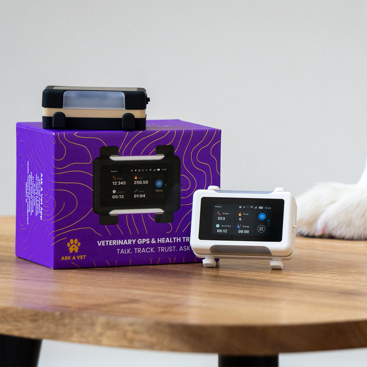 White / Gold | Smart GPS & Health Tracker: The White / Gold tracker beside its purple box on a table, with a dog's paw