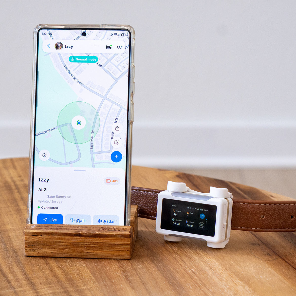 White / Gold | Smart GPS & Health Tracker: Phone in a wooden stand showing the live map, beside the White / Gold tracker on a collar